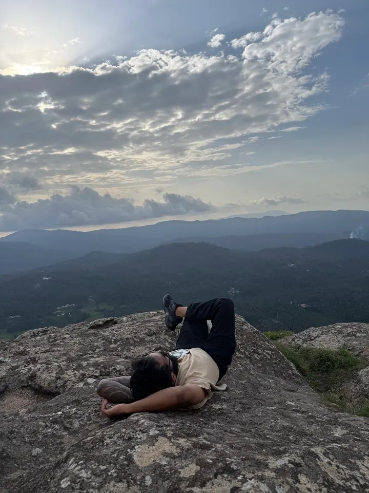 A trekker lying back on the summit rock of Bandekallu Gudda under a wide sky