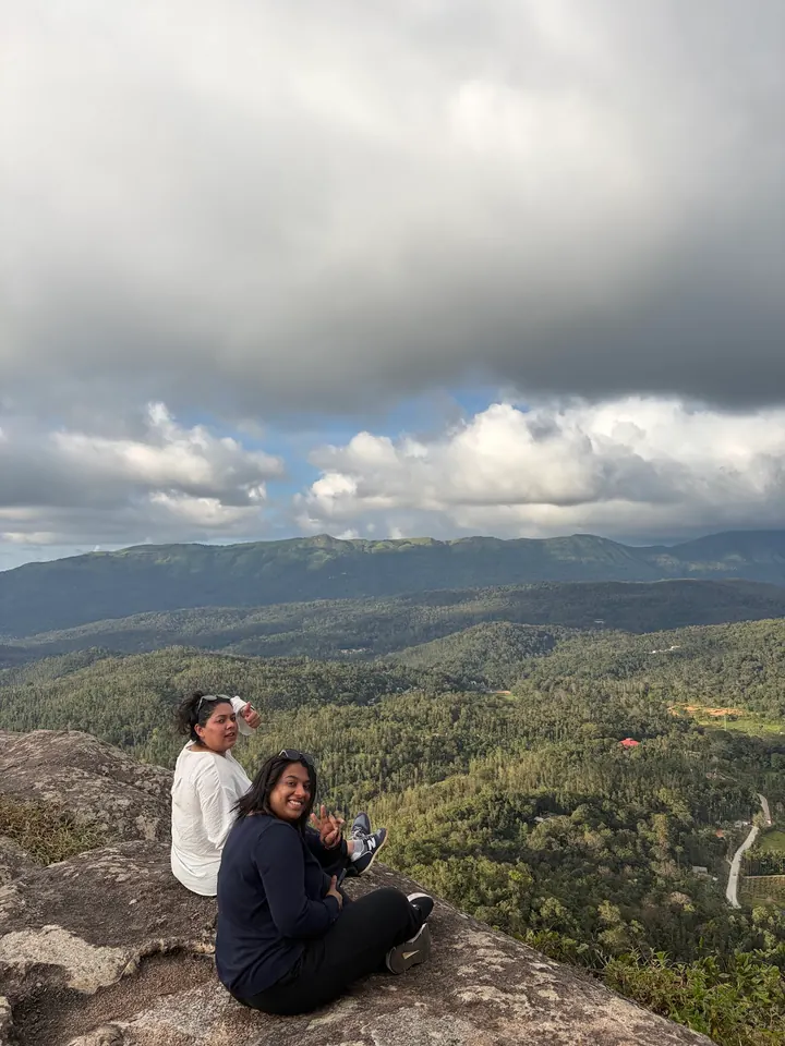Two friends smiling on the rocky top of Bandekallu Gudda, with green forest and hills behind them