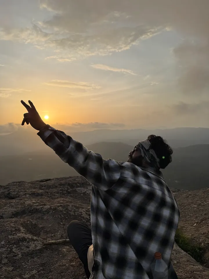 A trekker raising a hand towards the setting sun from the top of Bandekallu Gudda