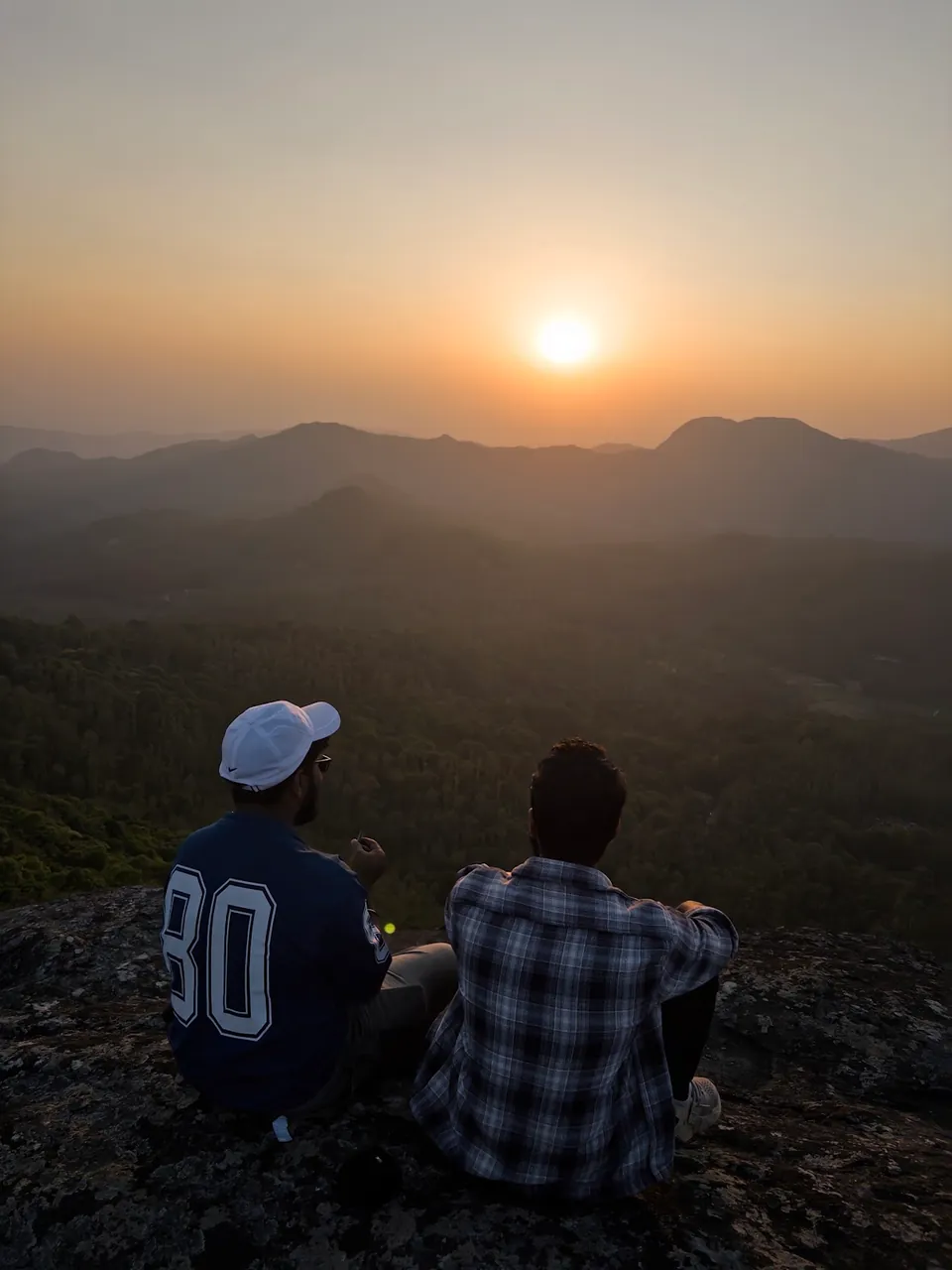 Two trekkers sitting on the rocky top of Bandekallu Gudda, watching the sun set over the hills